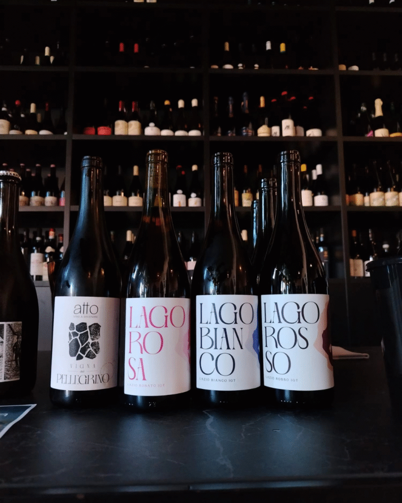 Five wine bottles lined up on a dark bar counter with a wall of bottles on shelves behind them.
