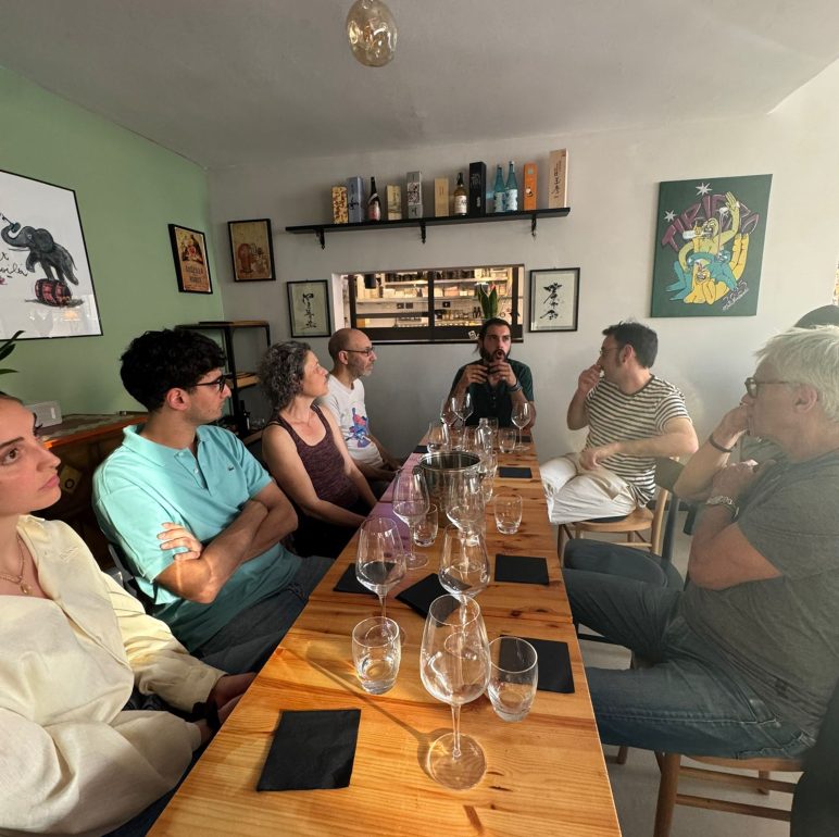 Group of people seated around a long wooden table in a cozy restaurant, wine glasses and black napkins in front of them.