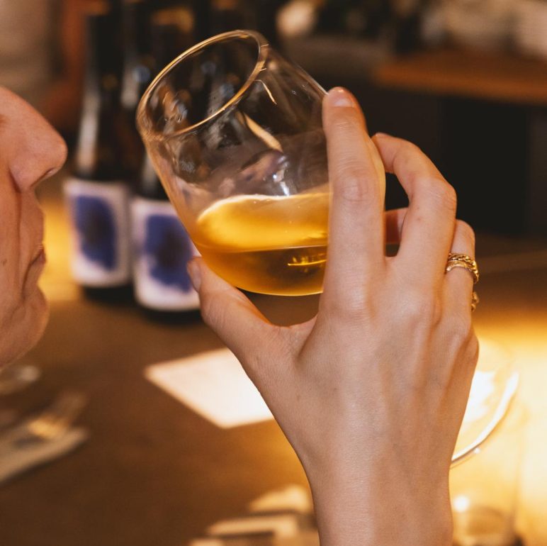 Close-up of a hand holding a glass of whiskey up to the mouth in a dim bar setting.