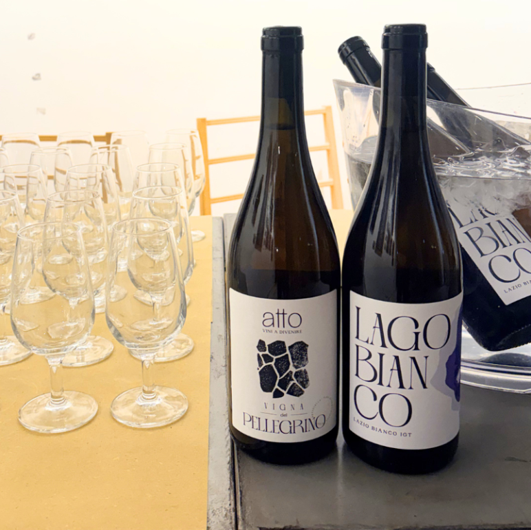 Two wine bottles (Pellegrino and Lago Bianco) stand on a table beside many empty wine glasses in a tasting setup.