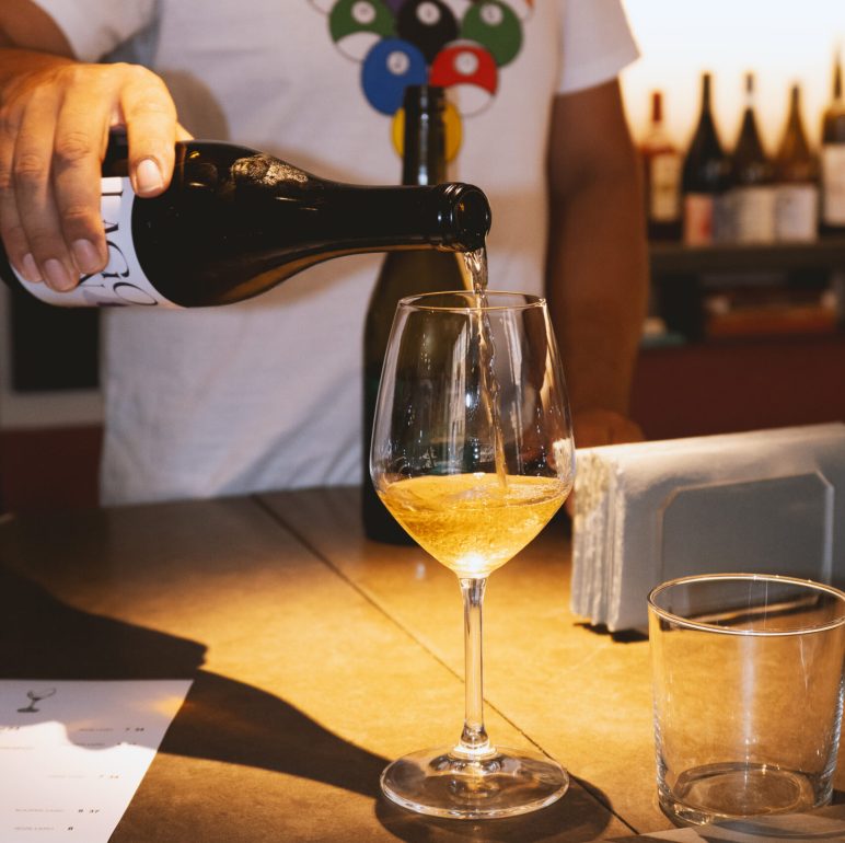Person pours sparkling wine from a dark bottle into a tall wine glass at a bar, with bottles on shelves in the background.