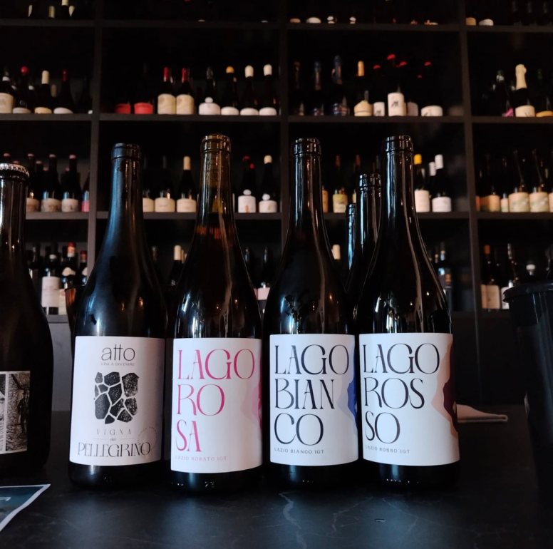 Five wine bottles lined up on a dark bar counter with a wall of bottles on shelves behind them.
