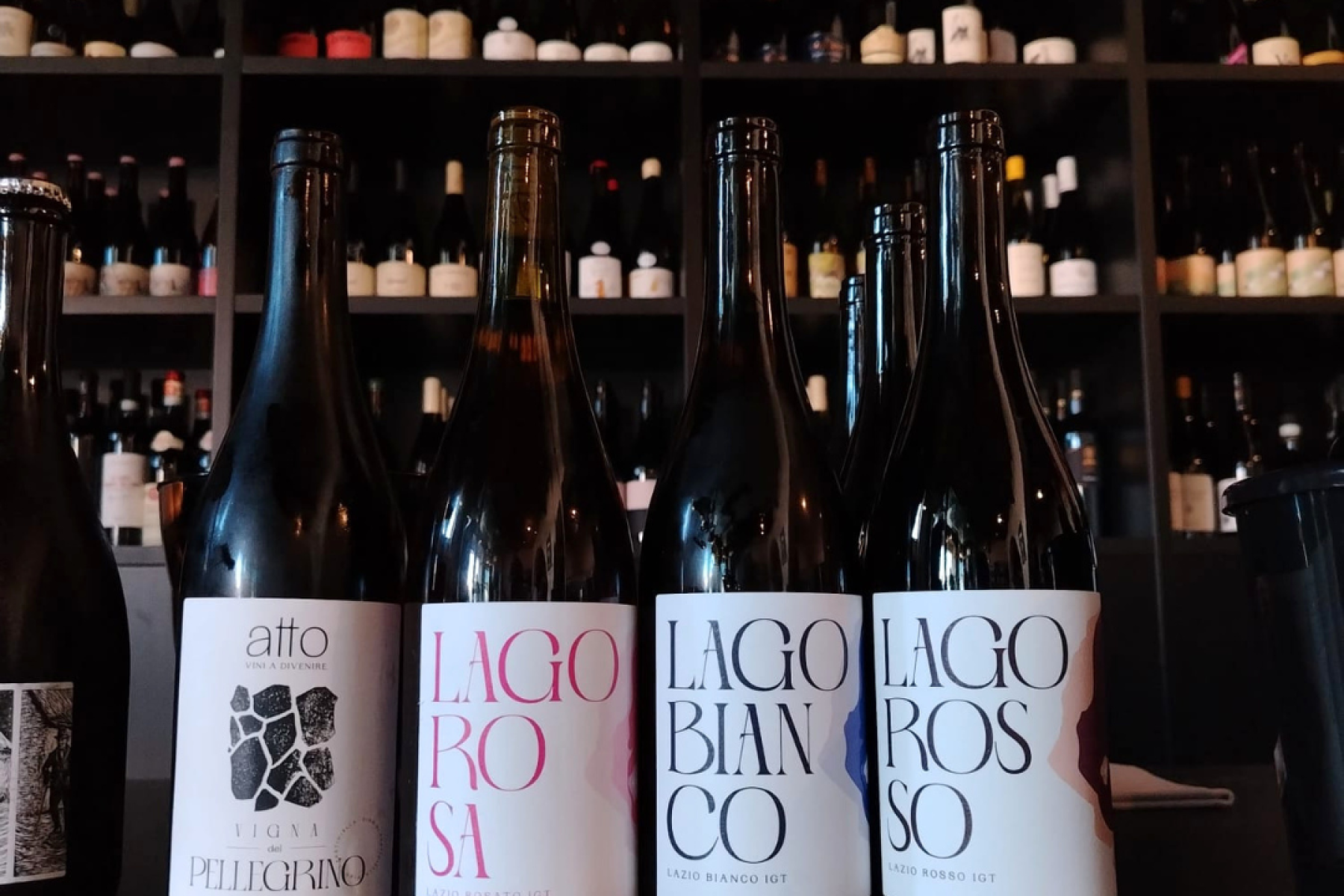 Five wine bottles lined up on a dark bar counter with a wall of bottles on shelves behind them.