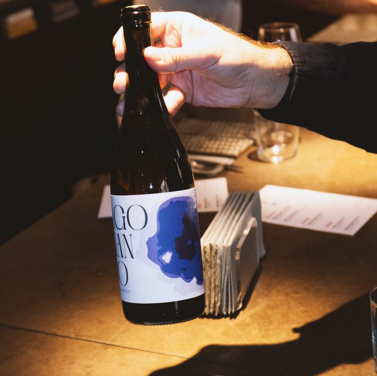 Person opening a dark beer bottle at a bar, with a blue-label bottle in focus.