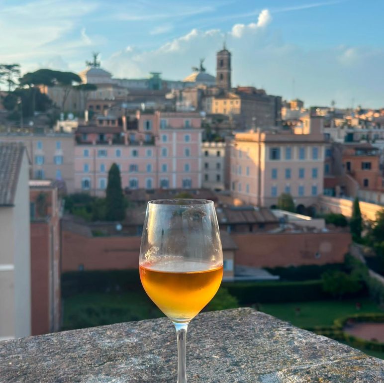 Wine glass with orange drink on a stone ledge overlooking a cityscape at sunset. Ca. warm light hits pastel buildings.